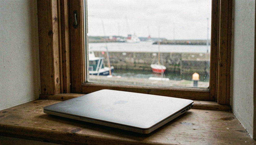 A closed laptop on a wooden windowsill looking out over a harbour wall