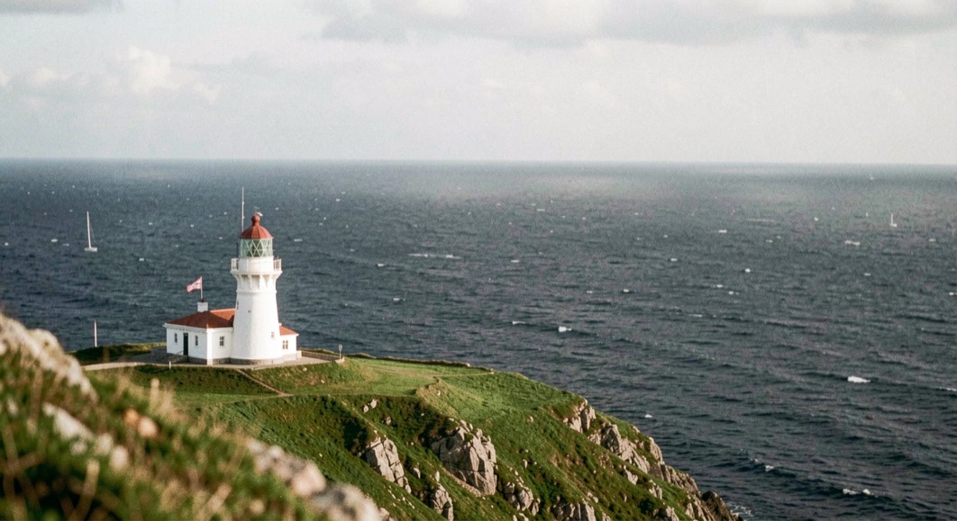 A white lighthouse on a low green headland above a dark sea with a long horizon behind